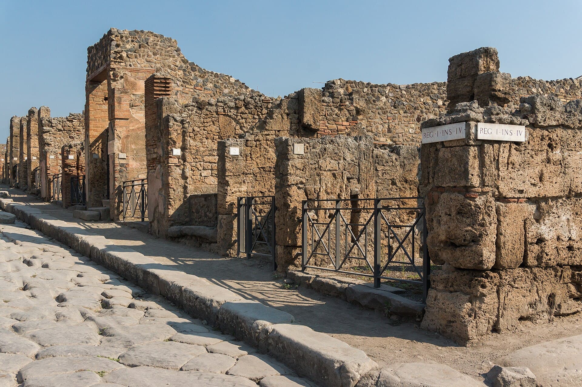 A cobbled street in Pompeii, lined with ruined stone walls, doorways, and a raised sidewalk.